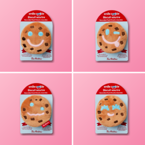 Pink promotional image featuring four Tim Hortons Smile Cookie reversible plush toys displayed in packaging. Each plush cookie has a different facial expression, including smiling, winking, confused, and laughing faces.