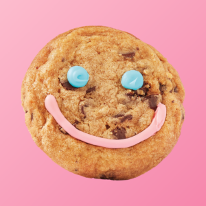 Close-up image of a Tim Hortons Smile Cookie on a pink background. The chocolate chip cookie is decorated with blue icing eyes and a pink icing smile.